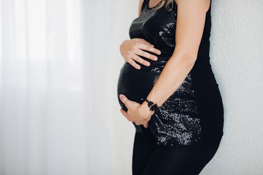 Close-up of unrecognizable pregnant woman in stylish black sparkling top embracing her tummy with two arms over white background. Copyspace.