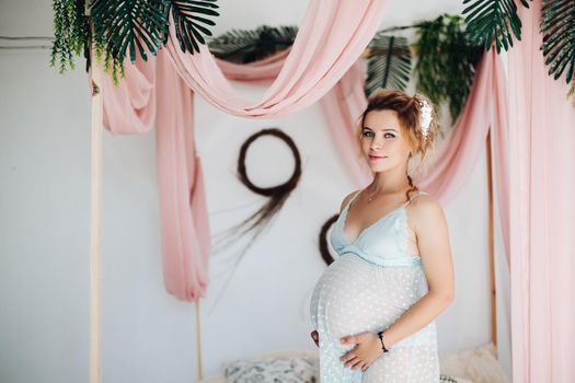 Portrait of pretty sensual pregnant lady in lace night gown embracing belly smiling at camera against decorated bed with bed curtains.
