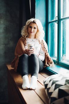 Portrait of pretty blonde woman in a woolen blanket, sitting next to blue colored window on windowsill. Beautiful lonely girl holding a cup in her hands. Concept of love and home.