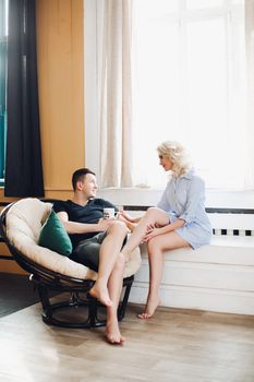 View of young beautiful couple against each other in stylish decorated interior near big window, looking in eyes each other. Family smiling and holding hands together, smiling, talking.