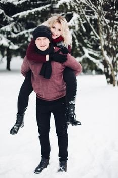 Happy couple posing, smiling and looking at camera in winter forest. Husband carrying his wife on back and beautiful blonde holding her hands on her significant other. Concept of winter hollidays.