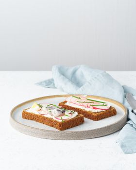 Savory smorrebrod, two traditional Danish sandwiches. Black rye bread with anchovy, radish, eggs, cream cheese on a grey plate on white stone table, vertical