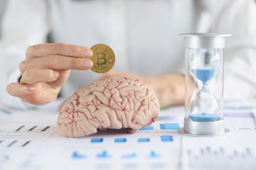 Close-up of businesswoman put bitcoin in human brain model, hourglass on table. Create idea, find ways to earn money, act fast. Intellect, invest concept