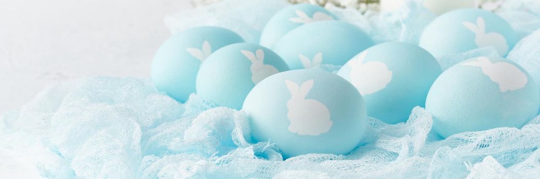 Easter banner. Holiday. Light white background, gentle pastel colors. Blue eggs with image of rabbit in basket. Flowers in background