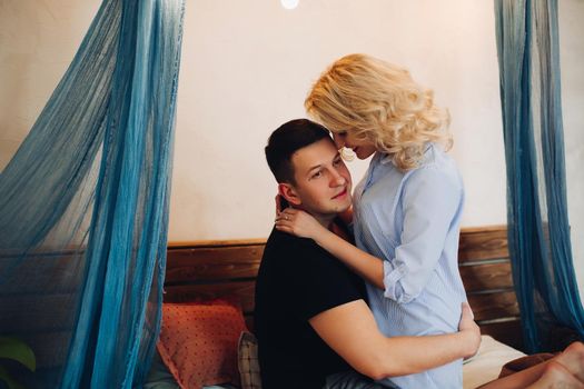 View of young happy couple sitting on bed in stylish bedroom and embracing each other. Beautiful blonde girlfriend with handsome boyfriend touching face by face. Home comfort.