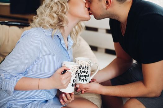 Close up of young sweet couple in love. Blonde girl in striped shirt and a man in black tshirt kissing tenderly,holding in their hands cups with creative inscription.Concept of love and relationship.