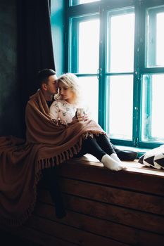 The couple wrapped in a woolen blanket, sitting and emracing next to the blue colored window on windowsill. Pretty blonde woman holding a cup in her hands.Concept of love and relationship,family.