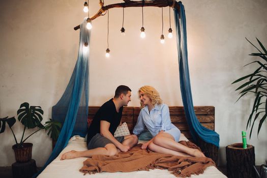 Young beautiful couple sitting together on bed in stylish decorated bedroom, looking in eyes each other. Family smiling and holding hands of each other, looking face to face.