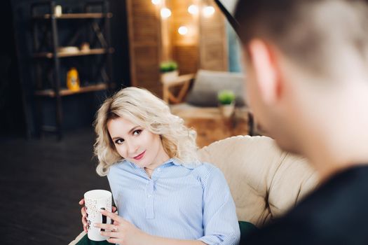 View of gorgeous blonde girlfriend sitting at coach against her boyfiend. Pretty wife looking eyes to eyes with his husband. Concept of love and relationship. Home interior.