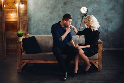 Portrait of romantic couple in love.Boyfriend with closed eyes tenderly kissing hand of his beautiful blonde girlfriend in black dress.They sitting on grey couch with black pillow in decorated studio.