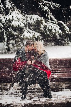 Amorous couple boyfriend and girlfriend posing and sitting on bench in winter forest. Pretty blonde and smiling man looking at camera and holding cups with coffee in hands. Concept of love.