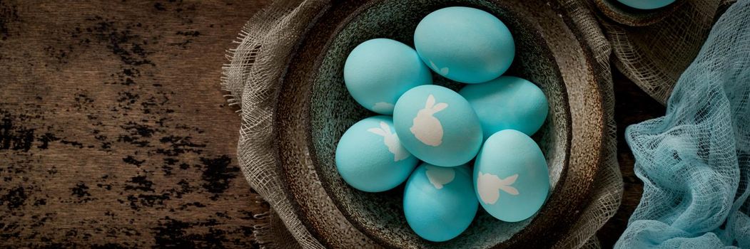 Unusual Easter on dark old background. Ceramic brown bowl with blue eggs with rabbit. Darkness, rays of sunlight shine on eggs. Concept of new life, rebirth. Rustic style. Top view, copy space