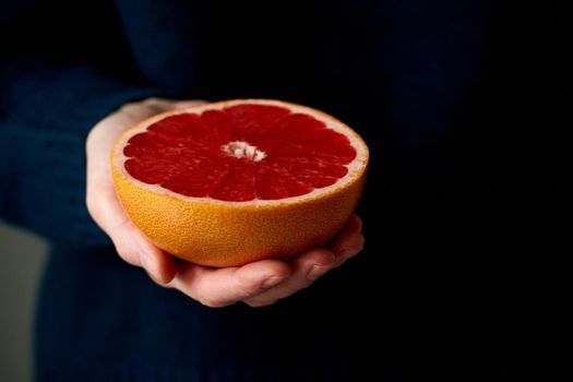 Close-up of woman's hand holding fresh halved red bright citrus fruit grapefruit on dark background. Lifestyle, natural light from window. Vitamins