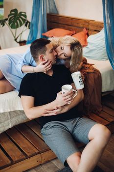 Young sweet couple in bed room, blonde girl in blue shirt and man in black t shirt kissing tenderly, holding in their hands cups with creative inscription.