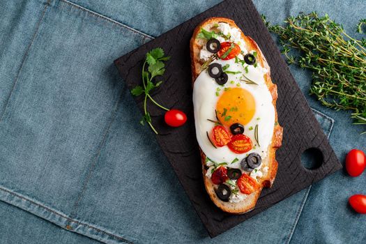 Toasted bread toast with fried eggs with yellow yolk and tomatoes, olives, sprinkled with herbs on dark wooden serving board on blue denim napkin, top view, copy space