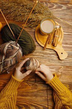 Women's hands with ball of yarn, coffee and cane on wooden background. Hands in orange sweater. Craft, hobby, top view. Wooden knitting needles in a ball of yarn. Cocoa and cinnamon sticks on a tray.