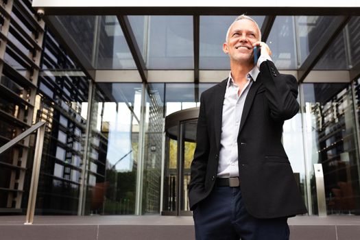Smiling caucasian businessman talking on mobile phone in front of office buildings. Copy space. Business and technology concept.