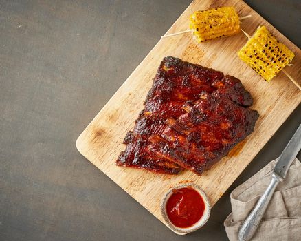 Barbecue pork ribs. Slow cooking recipe. Whole pickled roasted pork meat with red sauce and corn on cutting board, copy space, top view. Brown background