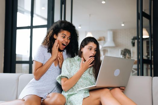 Two beautiful young women relaxing on the living room floor looking at a laptop computer and laughing at something on the screen