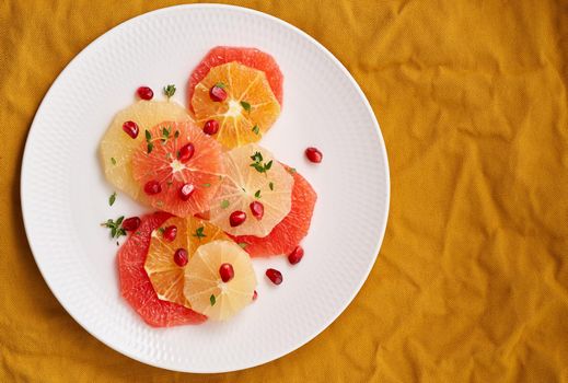 Bright mix of citrus fruits. Salad of mix sliced round slices of red and white grapefruit sprinkled with pomegranate seeds on white plate on yellow linen. Copy space