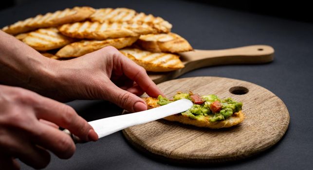 Bruschettas with toast, guacamole and cherry tomatoes on wooden board. Person hands prepare with knife mexican snack slice bread with avocado and vegetables
