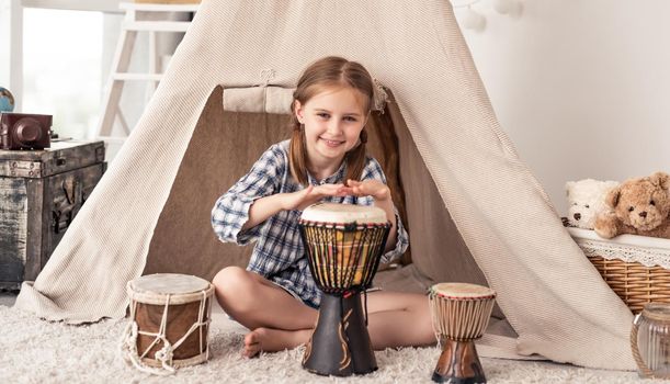 Little girl playing on traditional african djembe drums sitting in wigwam in kids room