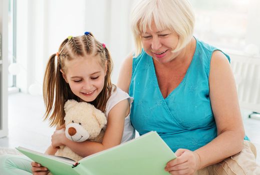 Smiling little girl reading book with happy elder woman indoors