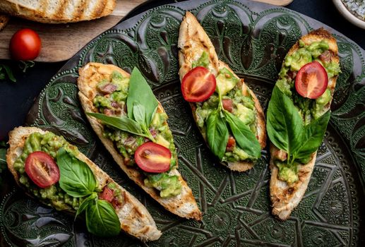 Bruschettas french toasts with guacamole, basil and cherry tomatoes served on beautiful plate with arugula herbs. Mexican snack antipasti with slice bread, mashed avocado and vegetables