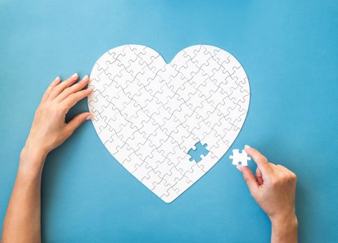 White puzzle in heart shape. Hands with white details of puzzle on blue background.