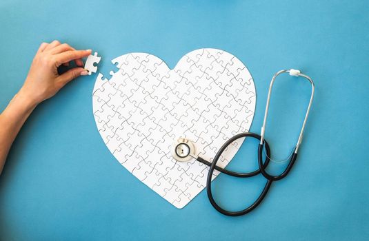 White puzzle in heart shape. Hands connect white details of puzzle on blue background. Heart health concept