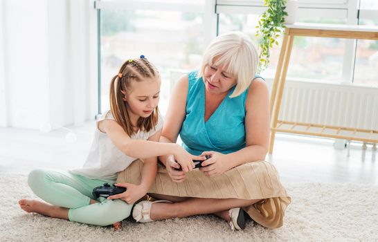 Cute little girl showing joystick buttons to granny sitting on floor in bright room
