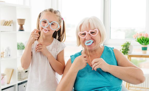 Smiling grandma having fun with cute granddaughter posing with paper glasses and lips at home