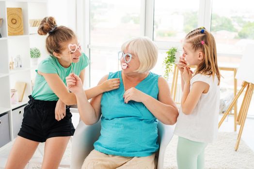 Cute granddaughters putting paper glasses and lips on smiling grandmother at home