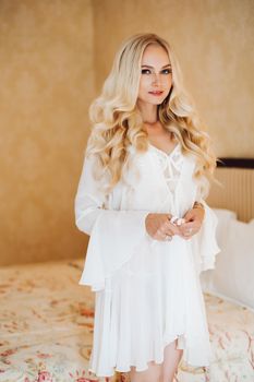 Sexy blondie bride in white lace lingerie and dress posing at luxury bedroom interior and undressing. Hot blonde haired woman holding dress by hand, looking away. Morrning of bride before wedding.