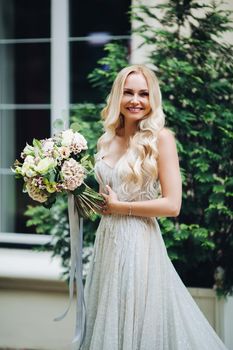 Front view of elegant and sensuality blondie bride looking at camera and smiling, holding wedding boquet. Beautiful female with makeup, wearing in wedding dress, posing outdoors.
