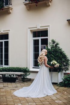 Back view of beautiful and sensuality bride in long, stylish wedding dress posing outdoors, against luxury house with wedding boquet. Beautiful wife waiting for her groom, sadly looking down.