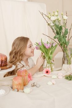 A girl with long hair in a light dress is sitting at the Easter table with cakes, spring flowers and quail eggs. Happy Easter celebration