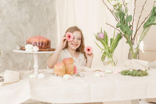 A girl with long hair in a light dress is sitting at the Easter table with cakes, spring flowers and quail eggs. Happy Easter celebration
