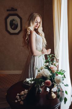 Portrait of beautiful and sensuality blondie bride sitting on chair in luxury interior, dreaming about husband, looking down. Young woman with long curly hair wearing in white lace lingerie.