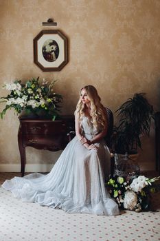 Gorgeous and beautiful blondie bride with long wavy hair, sitting on chair in luxury interior, looking at camera. Seductive woman wearing in white lace lingerie in morning. Wedding concept.