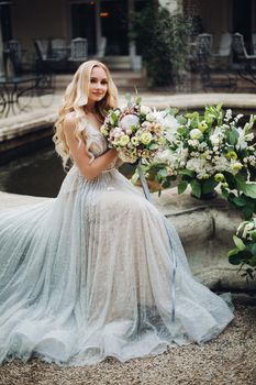 Picture of young seductive, elegant bride in wedding dress, sitting outdoor and posing at camera. Blond curly long hair attractive young girl holding flowers boquet in hand, looking down and smiling.