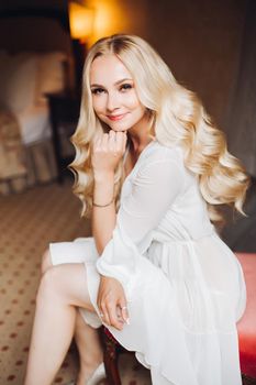 Beautiful and sensuality blondie bride sitting in bedroom, dreaming with closed eyes. Young and sexy woman with long curly hair and perfect make up, wearing in lace lingerie. Morning before wedding.