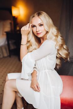 Beautiful and sensuality blondie bride sitting in bedroom, dreaming with closed eyes. Young and sexy woman with long curly hair and perfect make up, wearing in lace lingerie. Morning before wedding.