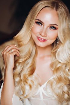 Portrait of young beautiful bride with wedding make up and hairstyle,wearing jewelry. Morning of bride.Happy long blondie hair girl in lingerie smiling, looking at camera.