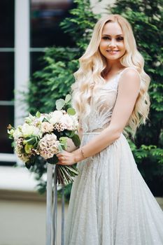 Front view of elegant and sensuality blondie bride looking at camera and smiling, holding wedding boquet. Beautiful female with makeup, wearing in wedding dress, posing outdoors.