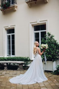 View of gorgeous and sensual bride in perfect, stylish wedding dress posing outdoors, against luxury house with wedding boquet. Beautiful wife waiting for her groom.