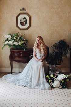 Gorgeous and beautiful blondie bride with long wavy hair, sitting on chair in luxury interior, looking at camera. Seductive woman wearing in white lace lingerie in morning. Wedding concept.