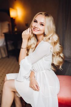 Beautiful and sensuality blondie bride sitting in bedroom, dreaming with closed eyes. Young and sexy woman with long curly hair and perfect make up, wearing in lace lingerie. Morning before wedding.