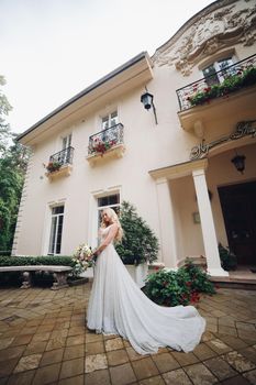 View of gorgeous and sensual bride in perfect, stylish wedding dress posing outdoors, against luxury house with wedding boquet. Beautiful wife waiting for her groom.