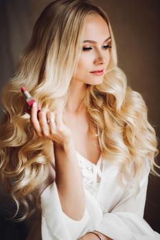 Beautiful and sensuality blondie bride sitting in bedroom, dreaming with closed eyes. Young and sexy woman with long curly hair and perfect make up, wearing in lace lingerie. Morning before wedding.